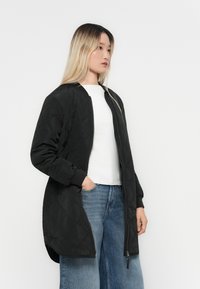 Black quilted jacket with a zipper, long sleeves, and ribbed cuffs. Worn over a white shirt and paired with light blue wide-leg jeans.