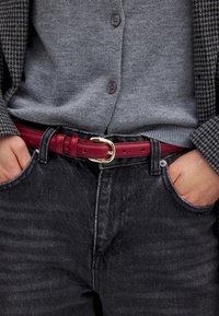 Červený kožený opasok so striebornou sponou nosený cez šedý kardigán a čierne denimové džínsy. Oblečenie má štruktúrovaný vzor a tmavé odtiene.