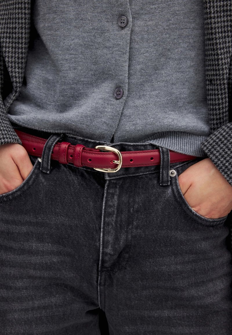 Červený kožený opasok so striebornou sponou nosený cez šedý kardigán a čierne denimové džínsy. Oblečenie má štruktúrovaný vzor a tmavé odtiene.