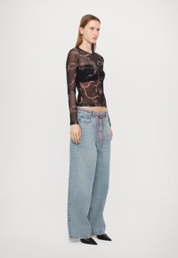 Schwarzes transparentes Langarmoberteil mit pinkem Blumendruck, kombiniert mit hellblauen weiten Jeans und buntem Kordelzug sowie schwarzen spitzen Schuhen.
