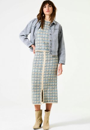 Femme portant une robe tricotée à motifs bleus et beiges avec une fente à l'avant, une veste en denim clair avec des clous, et des bottes à talons beiges sur fond blanc.
