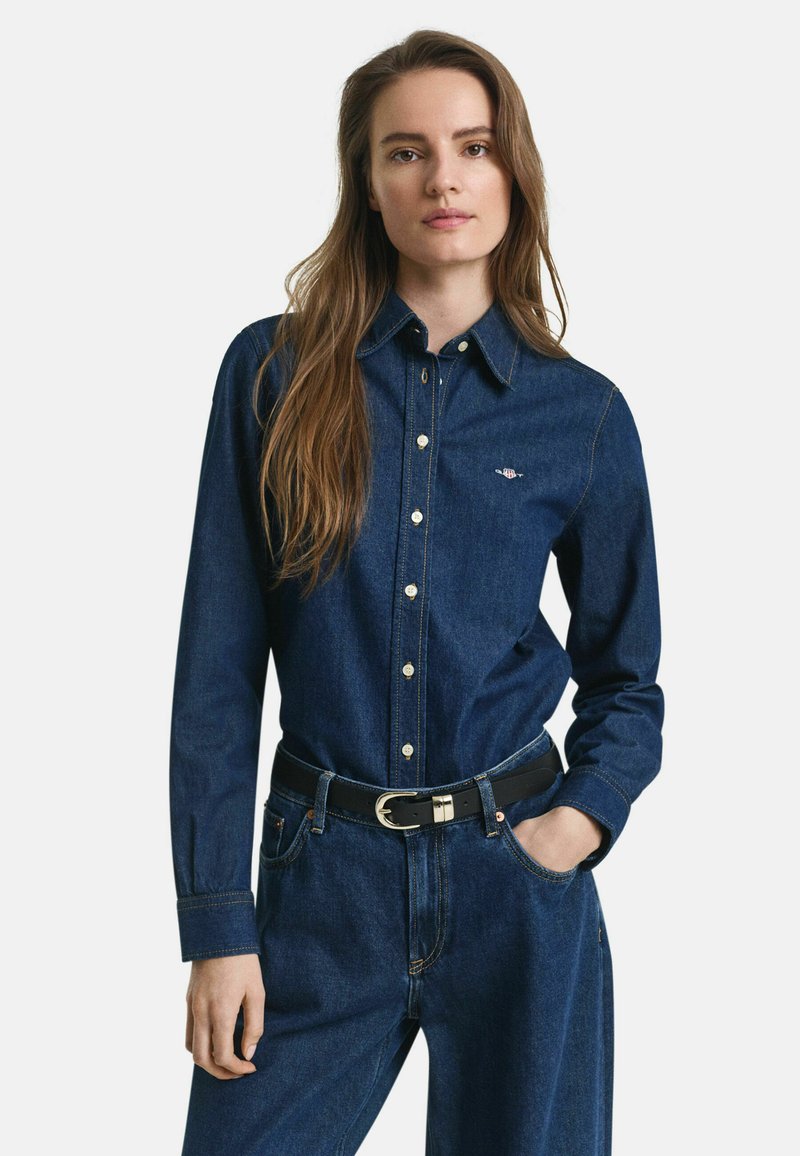 Woman with long brown hair wears buttoned dark blue denim shirt tucked into matching jeans with black belt, one hand in pocket.