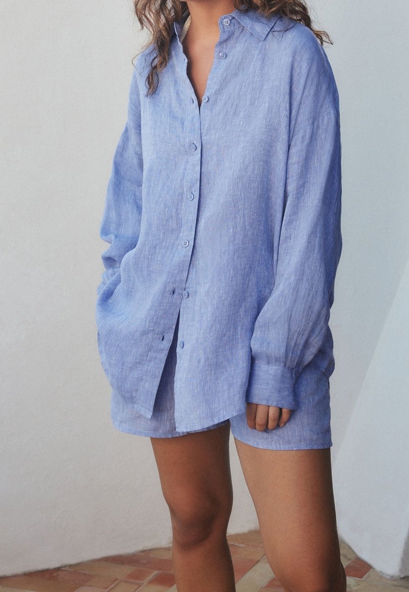 Femme portant une chemise bleu clair à manches longues boutonnée avec un short assorti, debout à l'intérieur sur un sol carrelé contre un mur blanc.