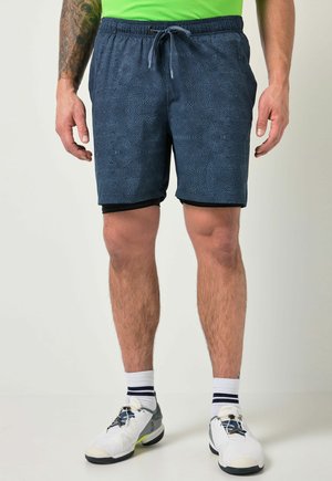 Mand iført mørkeblå teksturerede træningsshorts med sort inderfor, hvide sneakers, hvide sokker med sorte striber og en grøn skjorte.