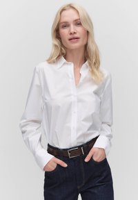Blondynka w białej koszuli zapinanej na guziki, włożonej w ciemne dżinsy z brązowym paskiem, stojąca z rękami w kieszeniach na jednolitym tle.