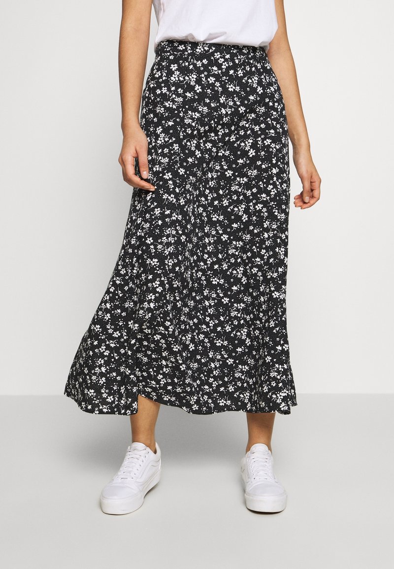 Personne portant une jupe midi noire imprimée de fleurs blanches, des baskets blanches et un haut blanc à manches courtes, debout devant un fond uni.