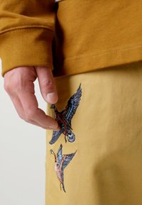 Gelbe Baumwollhosen mit detailliert gestickten Vögeln im Flug. Ein braunes Langarmhemd mit einem enganliegenden Bündchen ist teilweise sichtbar.
