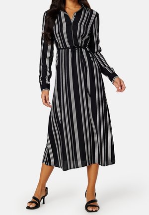 Femme portant une robe midi noire à rayures verticales blanches, manches longues, ceinture à la taille et sandales noires à talons.