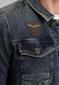 Veste en denim bleu foncé avec une texture usée, dotée d'un patch en bronze avec des ailes et de détails en boutons sur les poches de poitrine.