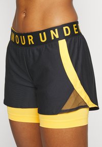 Svarta träningsshorts med en gul meshpanel och en integrerad gul innershorts. Midjebandet har texten "ARMOUR UNDER" i feta gula bokstäver.