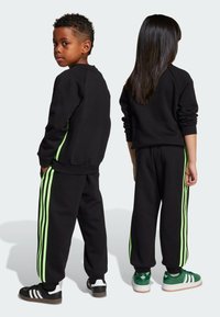 Schwarzer Trainingsanzug mit grünen Seitenstreifen, ausgestattet mit Rundhalsausschnitt und gerippten Bündchen. Zwei Kinder posieren Rücken an Rücken und zeigen das Design und die Passform.
