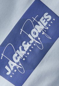 Το ανοιχτό μπλε ύφασμα έχει ένα εμφανές σκουρό μπλε ορθογώνιο πατς με λευκό κείμενο "JACK & JONES" και πάνω από αυτό μι α υπογραφή σε στυλ γραφής.