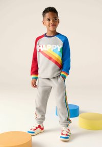 Šedá mikina s nápisom "HAPPY", červenými a modrými rukávmi a dúhovými pruhmi. Šedé tepláky s dúhovými bočnými akcentmi. Farebné topánky.