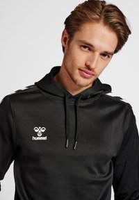 Felpa con cappuccio nera dal tessuto morbido, con logo bianco sul petto e accentuazioni bianche a contrasto sulle spalle. Cordini al cappuccio.