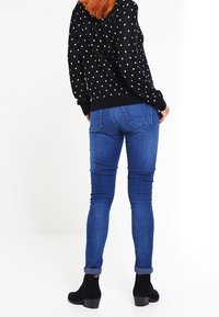 Czarny sweter w białe groszki, zestawiony z niebieskimi obcisłymi dżinsami. Model nosi czarne botki z fakturowanym wykończeniem.