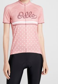 Koszulka rowerowa z krótkim rękawem w jasnoróżowym kolorze. Posiada białe kropki i paski, zamek błyskawiczny z przodu oraz logo marki w ciemniejszym odcieniu. Miękka tekstura.