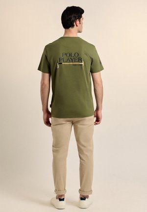 Camiseta verde oliva con el texto "JUGADOR DE POLO" en azul y un detalle de cremallera en la parte de atrás, combinada con pantalones beige y zapatillas blancas.