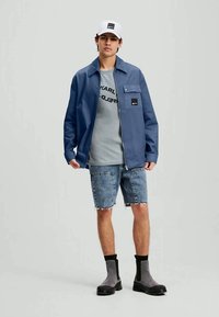 Jovem usando um boné branco, casaco azul com fecho, t-shirt cinza com logótipos, calções de ganga e botas cinzas, de pé contra um fundo simples.