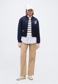 Femme portant une veste bomber bleu marine par-dessus un pull rayé et une chemise, un pantalon beige, des baskets argentées et une casquette de baseball blanche, posant devant un fond blanc.