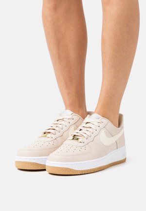 Beige Nike Air Force 1 Sneaker mit weißem Swoosh, weißen Zwischensohlen und gum-gummierten Außensohlen, getragen an nackten Beinen vor weißem Hintergrund.