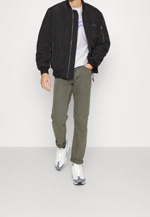 Schwarze Bomberjacke mit gerippten Bündchen, kombiniert mit olivgrünen Hosen und grauen Sportschuhen. Das Outfit beinhaltet ein weißes T-Shirt darunter.