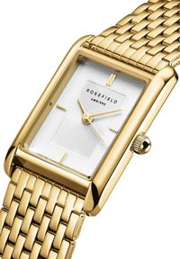 Montre rectangulaire dorée avec un cadran blanc texturé, des aiguilles dorées et un logo. Comprend un bracelet tressé doré et une couronne sur le côté.
