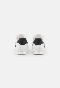 Hvite sneakers med en teksturert svart hælflik, glatte overflater, snøring og en merket sølvdetalj på hælen.