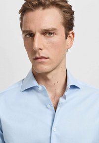 Svetlomodrá košeľa s golierom a vertikálnymi pruhmi, s gombíkovým zapínaním a hladkou textúrou. Tkanina pôsobí ľahko a sviežo.