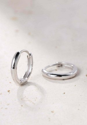 Srebrne kolczyki w formie obręczy o gładkim, zaokrąglonym wzornictwie. Wyposażone w zapięcie na zawias oraz minimalistyczną powierzchnię, podkreślającą refleksyjne wykończenie.