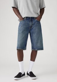Denim shorts in medium blue with a faded finish, featuring five pockets, and a straight cut. Paired with black slip-on shoes and white socks.