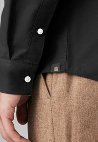 Cămașă neagră cu nasturi albi, mansetă detaliată și un tag mic cu sigla. Perechea este prezentată cu pantaloni texturați maro.