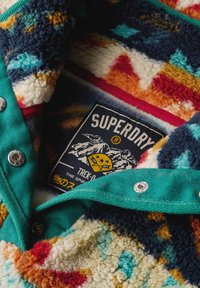 Gros plan d'une veste polaire colorée avec des boutons-pression métalliques et une étiquette Superdry Trek cousue à l'intérieur, illustrant des montagnes.