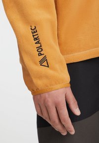 Fleece jakna v oranžni barvi z gladko teksturo. Ima črni Polartec logo na rokavu in elastični manšeti.