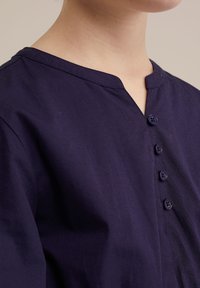 Mornarica bombažni srajc s V-izrezom in šestimi ujemajočimi gumbi ob zadrgi, ki ima gladko teksturo in rahlo sijaj.
