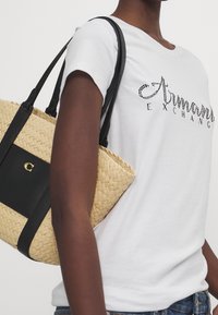 Bolso de paja tejido con detalles en cuero negro; cuenta con correas de hombro ajustables y una parte superior abierta. Modelo lleva una camiseta blanca con logo.
