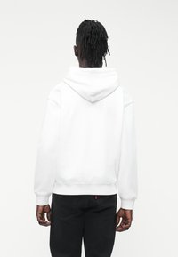 Pessoa com pele escura e dreadlocks, vestindo um capuz branco e calças pretas, de costas, diante de um fundo branco simples.