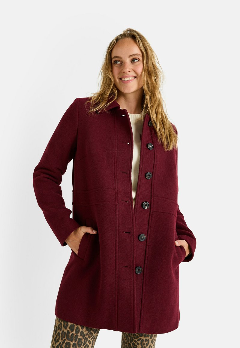 Abrigo de lana burdeos con cuello tipo solapa, cierre frontal de botones y bolsillos laterales. Presenta un corte entallado y una textura suave. Combinado con pantalones de estampado de leopardo.