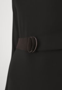 Černý látkový opasek s hladkým povrchem, zdobený kulatou kovovou sponou, upevněný kolem oděvu.