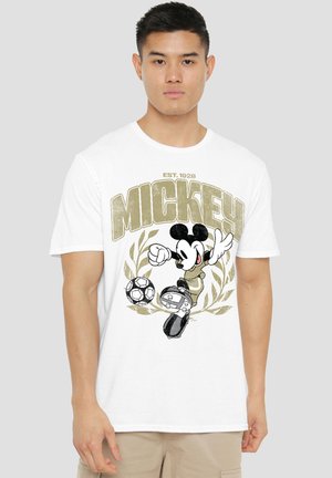 Junger Mann trägt ein weißes T-Shirt mit einem Mickey Mouse im Vintage-Stil, der einen Fußball kickt, und dem Text „MICKEY EST. 1928“.