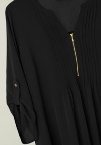 Sort bluse med plisseringer nær halsudskæringen, en halv-lynlås lukning foran og opkrøllede ærmer med knapdetaljer i guld-tone. Glat stoftekstur.