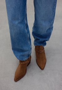 Brown suede ankle boots with pointed toes, embellished with silver studs on ankle straps, paired with loose blue denim jeans.