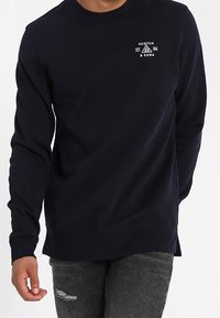 Sudadera de cuello redondo de la marina, confeccionada en una tela suave con puños acanalados. Presenta un logo blanco en el pecho y un corte relajado, acompañada de jeans desgastados.