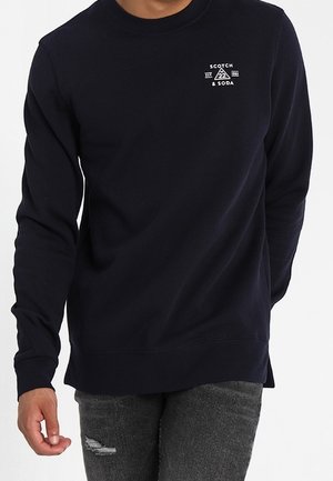 Sudadera de cuello redondo de la marina, confeccionada en una tela suave con puños acanalados. Presenta un logo blanco en el pecho y un corte relajado, acompañada de jeans desgastados.