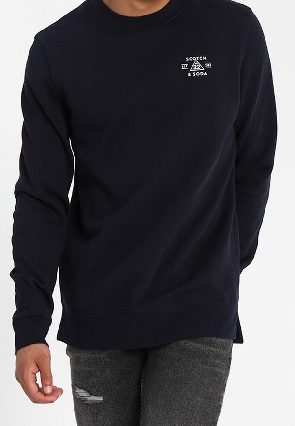 Sudadera de cuello redondo de la marina, confeccionada en una tela suave con puños acanalados. Presenta un logo blanco en el pecho y un corte relajado, acompañada de jeans desgastados.