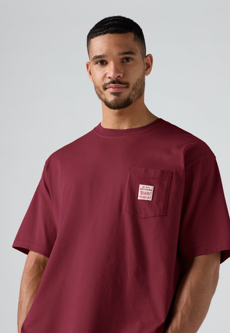 Uomo con capelli corti e barba che indossa una maglietta larga color bordeaux con una piccola toppa bianca sulla tasca del petto su uno sfondo chiaro e uniforme.