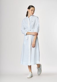 Abito midi in cotone bianco con vita regolata da una cintura, collo alto e maniche a sbuffo. Abbinato a sneakers bianche, evidenziando un design pulito e minimalista.