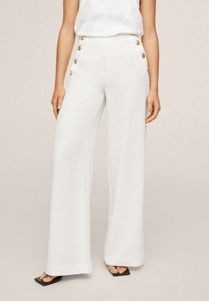 Kvinde iført hvide vidde-bukser med høj talje og guldknapper i siderne, kombineret med en hvid ærmeløs top og sorte sandaler med firkantet tå.