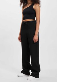 Black ribbed crop top with one shoulder strap, paired with loose-fitting black trousers and white sneakers. Smooth texture, relaxed fit.