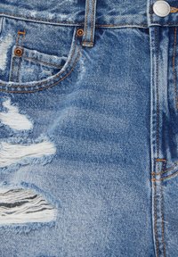 Denim azul claro desgastado con bordes deshilachados y costuras naranjas en contraste, con bolsillos visibles y un botón de metal.