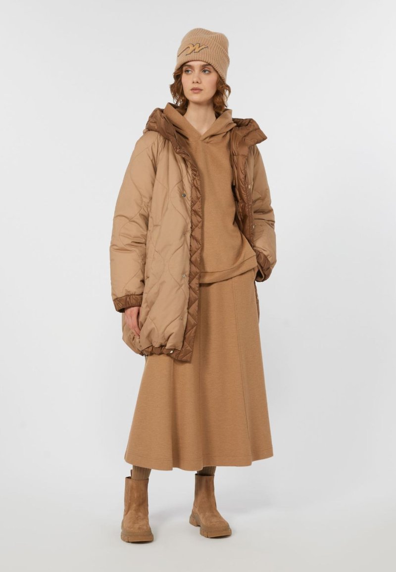 Frau in einer beigen Strickmütze, braunem Kapuzenpullover, gepolsterter beige Jacke, langem beigen Rock und beigen Stiefeletten, die vor einem schlichten weißen Hintergrund steht.